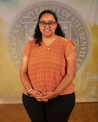 Gaby Taylor in an orange blouse standing in front of the university seal.