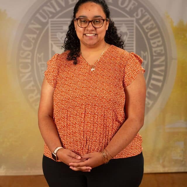 Gaby Taylor in an orange blouse standing in front of the university seal.