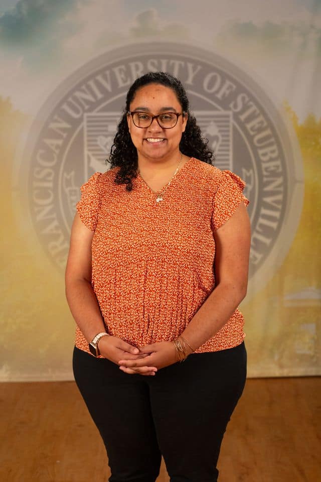 Gaby Taylor in an orange blouse standing in front of the university seal.