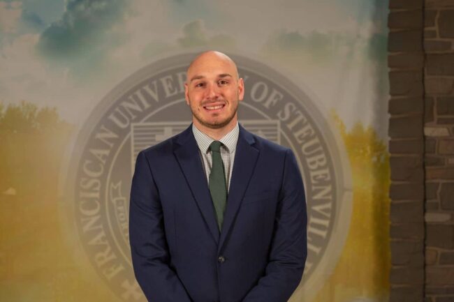 Aaron Foldi in a suit standing in front of a university seal.
