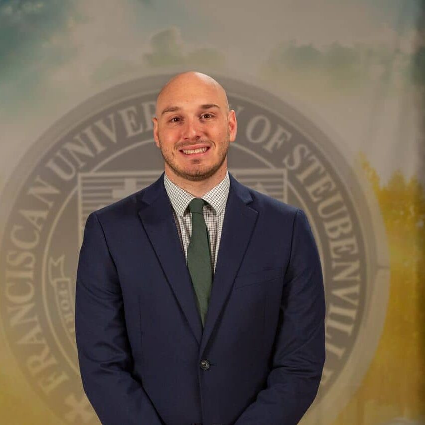 Aaron Foldi in a suit standing in front of a university seal.