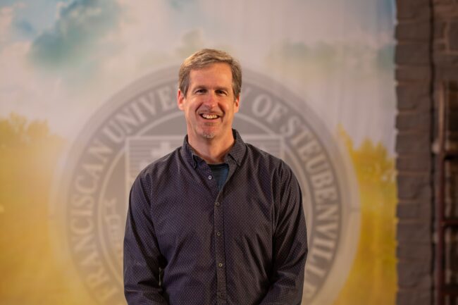 Dr. Dan Kuebler standing in front of the Franciscan University of Steubenville seal.