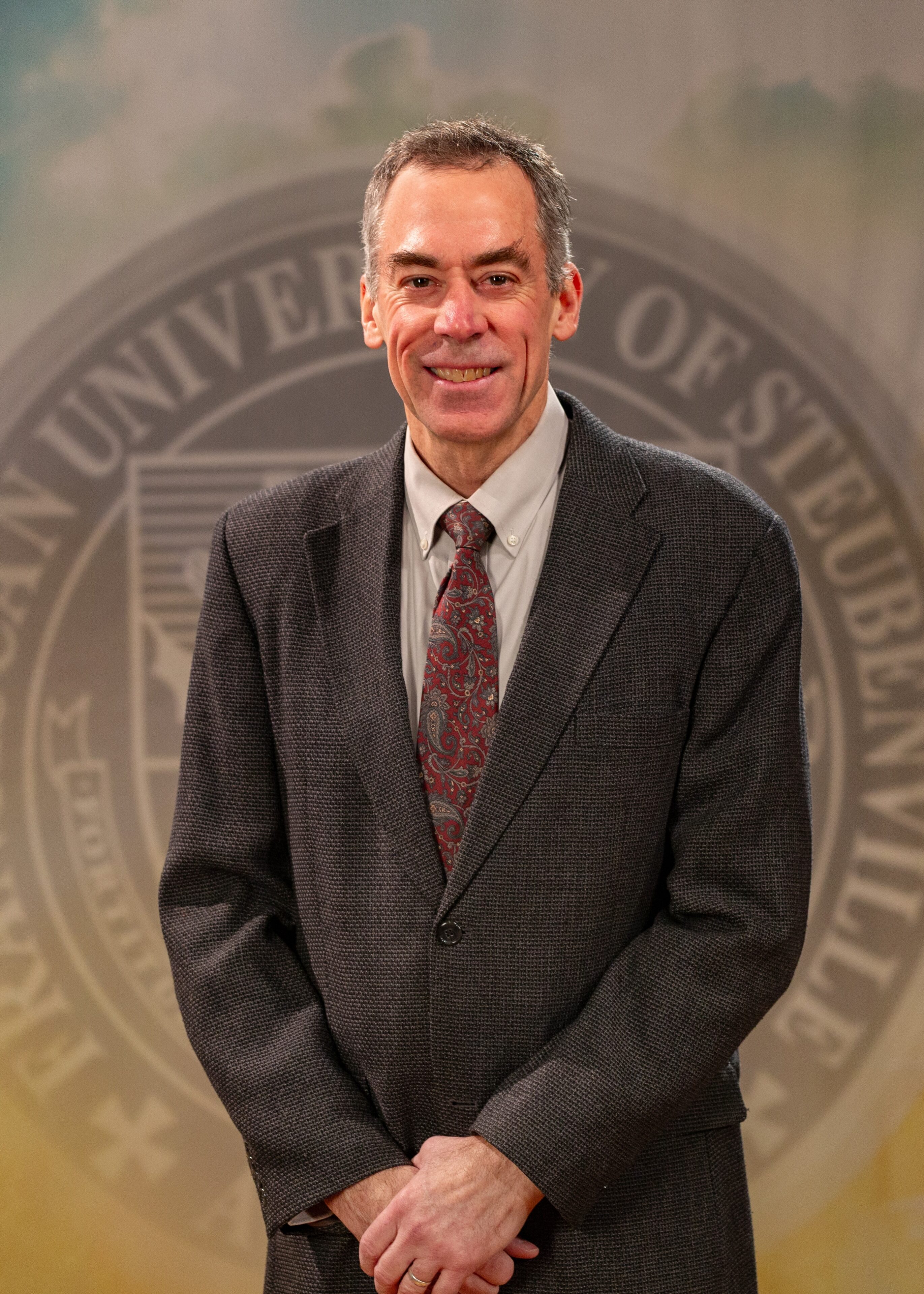 Dr. Ron Bolster_Faculty Portrait_Spring2026_-2816 Portrait of Dr. Ron Bolster standing in front of the Franciscan University of Steubenville seal.