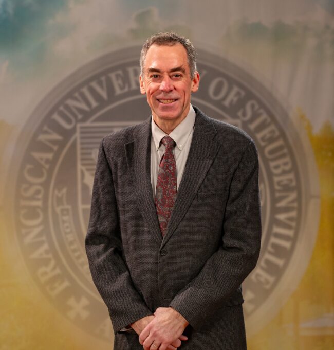 Portrait of Dr. Ron Bolster standing in front of the Franciscan University of Steubenville seal.